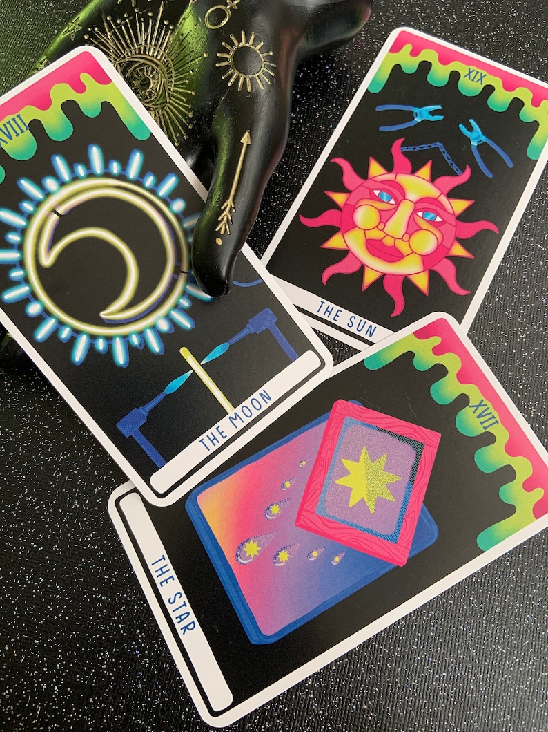 May include: Three tarot cards with a neon-coloured design. The cards are The Moon, The Star, and The Sun. The Moon card features a crescent moon with a glowing halo, The Star card shows a star in a frame with shooting stars, and The Sun card depicts a smiling sun with a chain and pliers.