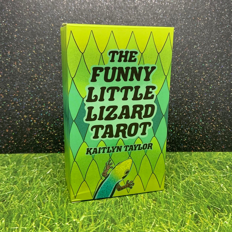 May include: A green and yellow box with a green lizard on the front. The box says "The Funny Little Lizard Tarot" and "Kaitlyn Taylor" in black text.