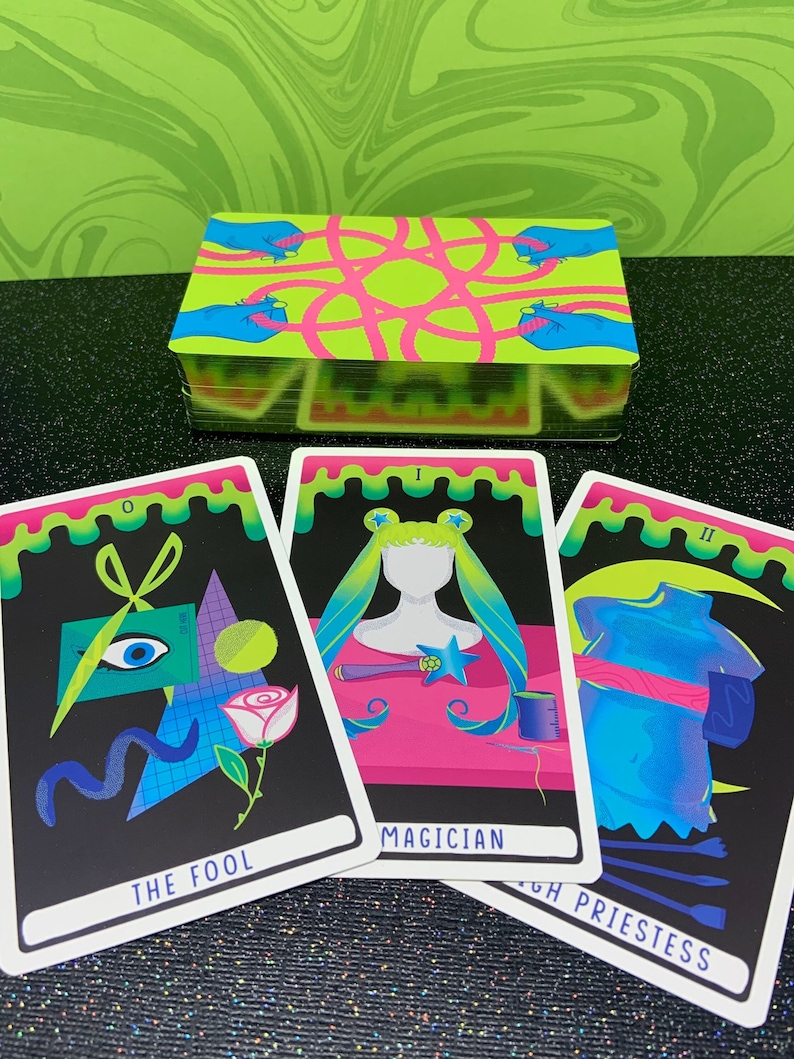 May include: Three tarot cards with neon colours and dripping edges. The Fool card shows a black background with a blue eye, a white rose, a pair of scissors, and a blue wavy line. The Magician card shows a pink background with a figure wearing a blue and green wig, a pink dress, and a star on their chest. The High Priestess card shows a blue background with a figure wearing a blue dress with a pink sash, and a crescent moon. The cards are labelled with their names and Roman numerals.