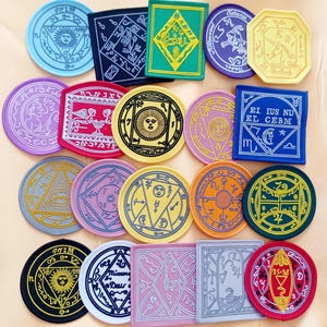 May include: A collection of 16 embroidered patches featuring various symbols, designs, and text. The patches are circular, square, and octagonal in shape and come in a variety of colours, including red, blue, green, yellow, and grey. The patches feature intricate designs and text, including the words "EI IUS NU EL CEBM" and "Micrors Deur".