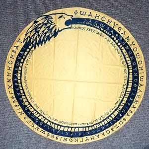 May include: A round, yellow tapestry with a black and blue design. The design features a stylized bird head with a long beak and a circle of Greek letters around the edge. The letters are in black and are arranged in a circular pattern.
