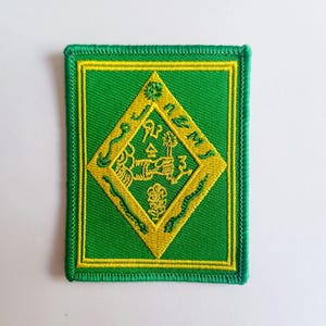 May include: Green and yellow embroidered patch with a diamond-shaped design. The diamond features a stylised figure holding a flower and surrounded by snakes. The text "NUMS" is written around the diamond.