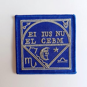 May include: A blue embroidered patch with silver thread. The patch features a circle with the text "EI IUS NU EL CEBM" inside. The circle is surrounded by a square with astrological symbols in the corners.