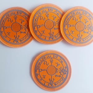 May include: Four orange round embroidered patches with a black circular design featuring a symbol with four arms and a circle in the center. The symbol is surrounded by a circle of text.