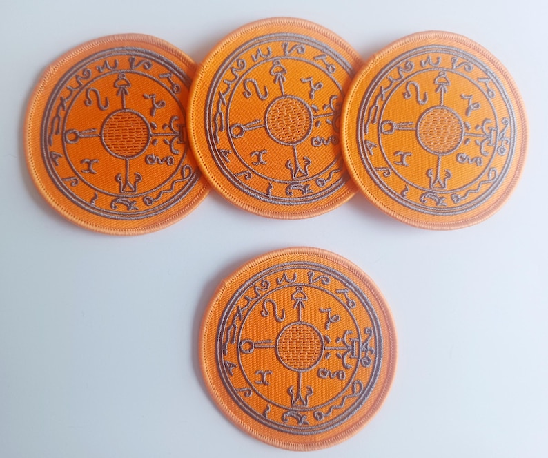May include: Four orange round embroidered patches with a black circular design featuring a symbol with four arms and a circle in the center. The symbol is surrounded by a circle of text.