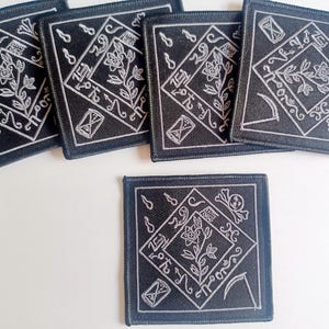 May include: Five black square patches with white embroidered designs. Each patch features a stylized flower with a stem and leaves, a crescent moon, a scythe, a skull, and an hourglass. The patches are approximately 4 inches by 4 inches.
