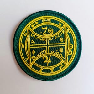 May include: A green and yellow embroidered patch with a circular design. The centre of the patch features the numbers "7" and "24" with a symbol resembling an eye. The patch has a decorative border with a repeating pattern.