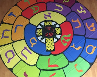 May include: A colorful circular rug with a central cross design and Hebrew letters around the perimeter. The rug is made of fabric and has a variety of colors, including red, yellow, green, blue, and purple.