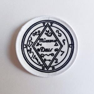 May include: White embroidered patch with a black circle and a diamond shape inside. The diamond shape has the words "Minacce" and "Deus" inside. The circle has various symbols and numbers around the edge.