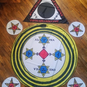 May include: A yellow and green circular cloth with a black snake design and a white center featuring four six-pointed stars with a red diamond in the center. The stars have blue and black accents. The text "ALPHA", "OMEGA", "TETRAGRAMMATON", "PRIMEUMATON", and "NAPHAXETON" are written around the outer edge of the cloth. Four white circular cloths with a six-pointed star with red and yellow accents are placed around the circular cloth.
