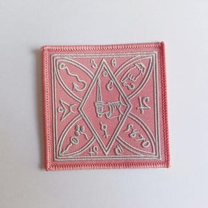 May include: A pink embroidered patch featuring a white design of a diamond shape with various symbols inside. The symbols include a sword, a hand, a bird, and a fish.