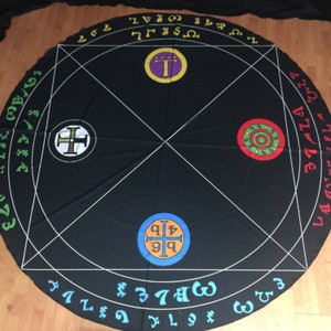 May include: A black circular cloth with white lines forming a geometric pattern. The cloth has four circular symbols in the center, each with a different color and design. The symbols are surrounded by a circle of numbers and letters.