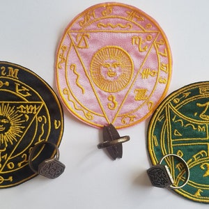 Puede incluir: Tres parches bordados con diseños intrincados. Uno es negro con dorado, otro rosa con dorado y el tercero verde con dorado. Cada parche tiene un anillo adjunto. Los parches presentan símbolos solares y geométricos.