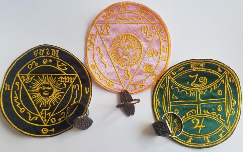 May include: Three embroidered patches with intricate designs. One is black with gold, another is pink with gold, and the third is green with gold. Each patch has a ring attached. The patches feature sun and geometric symbols.