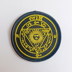 以下が含まれることがあります： 黒と金の刺繍が施されたパッチで、円形のデザインになっています。円の真ん中には、太陽の顔が入った三角形があります。円は「M2SM」という文字の輪で囲まれています。