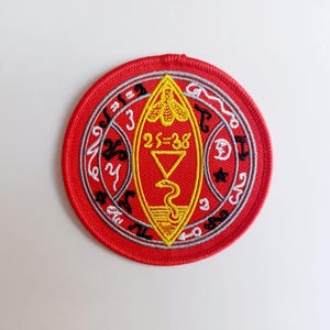 May include: Red embroidered patch featuring a yellow and black symbol within a circle. The symbol depicts a snake, a triangle, and the text "25=38". The circle has a grey and white design with black text.