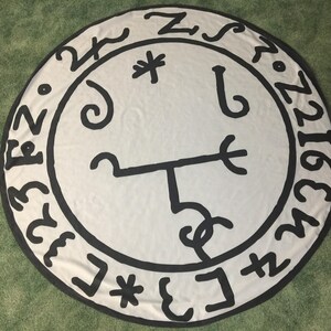 May include: A white circular rug with a black sigil in the center. The sigil is surrounded by a circle of numbers and symbols. The rug is on a green surface.