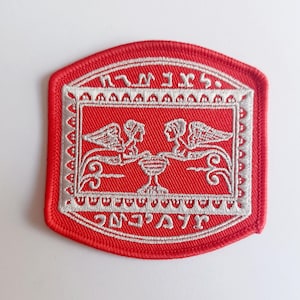 May include: Red embroidered patch with a white design of two winged figures holding a chalice. The text "אמת ואהבה" is written in Hebrew around the border.