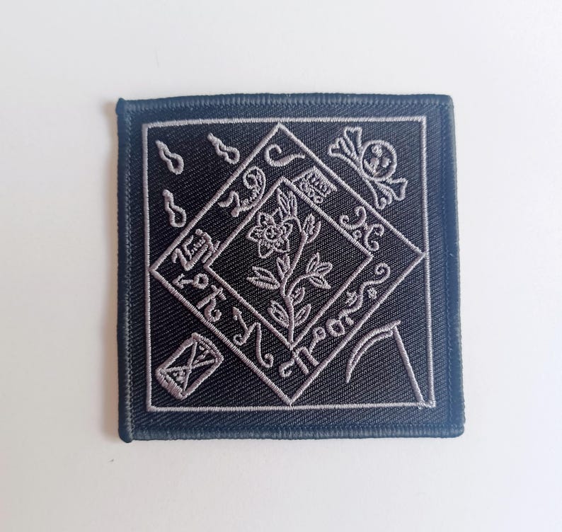 May include: Black embroidered patch with a silver floral design inside a diamond shape. The design includes a skull and crossbones, a scythe, and other symbols.