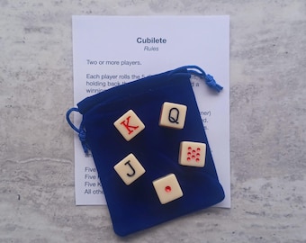 May include: A blue velvet drawstring bag containing five ivory-colored dice with red and black markings. The dice show the letters K, Q, and J, and the numbers 1 and 5. A white paper with the game rules is behind the bag.