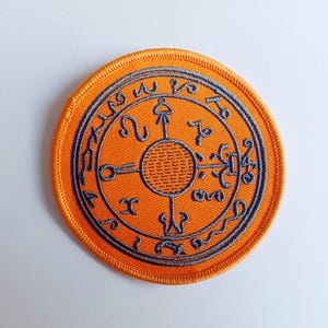 以下が含まれることがあります： オレンジ色の刺繍パッチで、円形のデザインに、中央に円形のグレーのシンボル、周囲にテキストが配置されています。