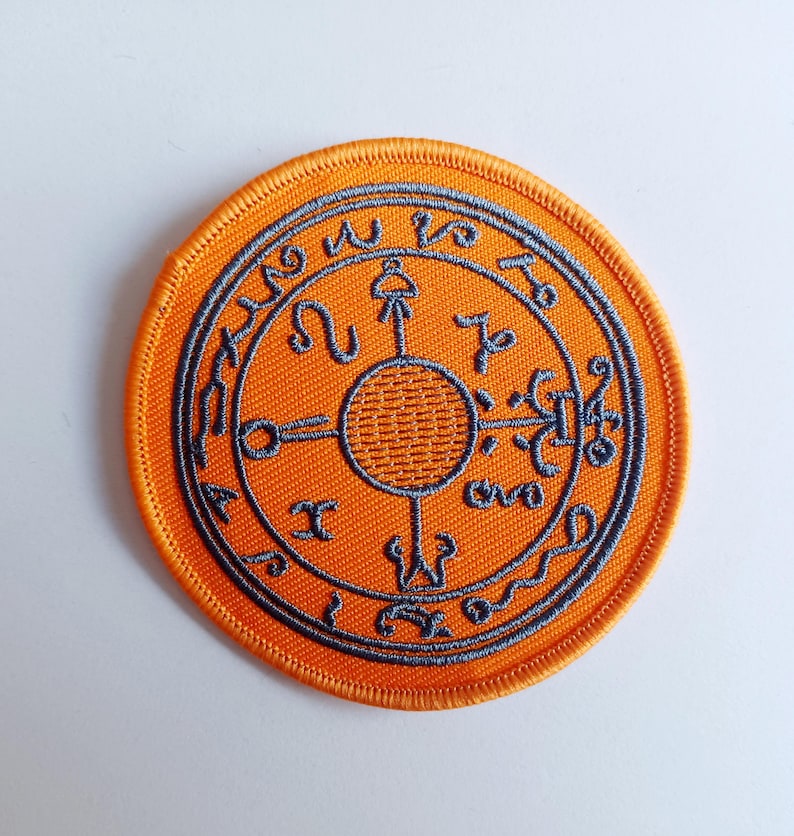 May include: Orange embroidered patch with a circular design featuring a gray symbol with a circle in the center and text around the outside.