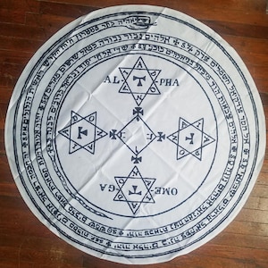 May include: A white circular cloth with a black and white design. The design features a circle with a star and a cross in the center. The circle is surrounded by a ring of text in Hebrew. The text reads "AL-PHA" and "OME-GA".