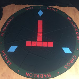 May include: A black circular cloth with a green border and red and blue symbols. The text around the border reads "PERDVRABO", "NVIT", "HADIT", "RAHOOR KHVI", "CHAOS", "BABALON", "L'VAYAH".