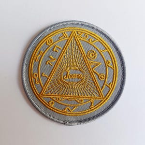 May include: A circular, grey embroidered patch with a gold design. The design includes a triangle with radiating lines, encircled by symbols and the word "Ieova". The patch has a stitched border.