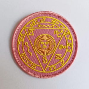 May include: A pink round embroidered patch with a yellow sun and a triangle with the number 3 inside. The triangle is surrounded by a circle of yellow embroidered symbols.