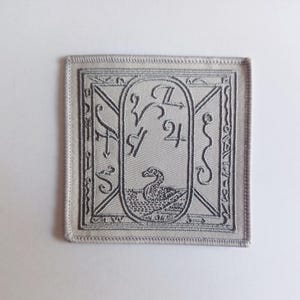 May include: A gray embroidered patch with a white background. The patch features a stylized swan with a number 24 and other symbols.