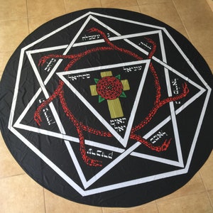 May include: A black circular tapestry with a white eight-pointed star design. The star is filled with a red snake design and Hebrew text. In the center of the star is a gold cross with a red rose.