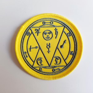 May include: A yellow embroidered patch with a grey circle and a grey triangle inside. The triangle has symbols and a face inside.