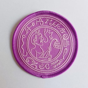 May include: Purple embroidered patch with a white outline. The patch features a stylised image of a person holding a flower. The text "I AM" is written around the image.