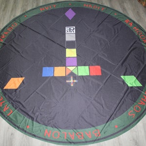 Puede incluir: Un paño circular negro con un borde verde y texto rojo. El texto dice "BABALON", "CHAOS", "L.A.W.M.I", "PERDURABO", "HADIT", "NVIT", "RAH-HOOR-KHVIT". El centro del paño presenta un símbolo en blanco y negro con un cuadrado gris, un cuadrado amarillo, un cuadrado morado y un cuadrado verde. El símbolo está rodeado por un cuadrado rojo, un cuadrado verde, un cuadrado azul y un cuadrado naranja.