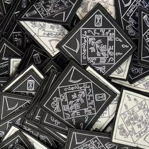 May include: A pile of black and white embroidered patches with a variety of designs, including a crescent moon, a flower, and a skull. The patches are all diamond-shaped and have a white outline.