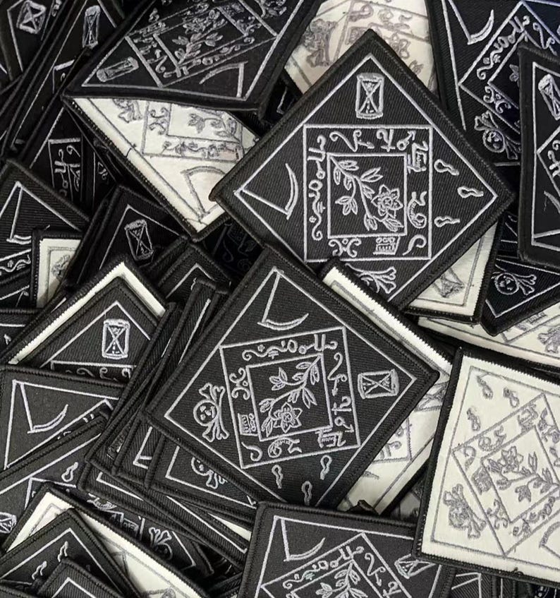 May include: A pile of black and white embroidered patches with a variety of designs, including a crescent moon, a flower, and a skull. The patches are all diamond-shaped and have a white outline.