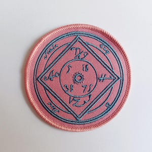 May include: A pink round embroidered patch with a blue circle and a blue diamond inside. The circle has numbers and symbols inside. The diamond has symbols and numbers around the edges.