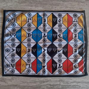 Puede incluir: Un tablero de juego de tela blanca con un patrón geométrico de cuadrados negros, amarillos, azules y rojos. Cada cuadrado contiene un símbolo, incluidos círculos, triángulos y letras.