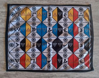 May include: A white fabric game board with a geometric pattern of black, yellow, blue, and red squares. Each square contains a symbol, including circles, triangles, and letters.