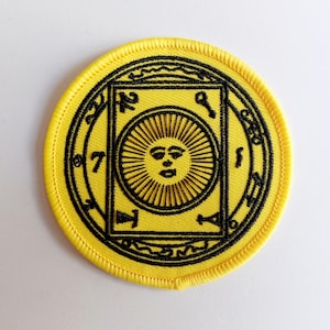 May include: Yellow embroidered patch with a black circle around a yellow square with a black sun inside. The sun has a face and rays. The circle has black text around the outside.