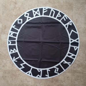 May include: A black round mat with a white geometric border. The border features stylized runes or symbols. The mat is made of a smooth fabric and is laid out on a light-colored tiled surface. The design is symmetrical.