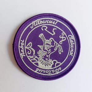 May include: Purple embroidered patch with a white design of a creature with a crown and the text "J'accuse!" and "Ritual 25 Classical"