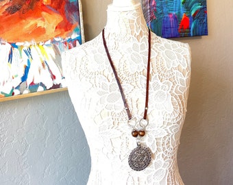 May include: A brown leather cord necklace with a silver pendant featuring an intricate design. The pendant is attached to a silver ring with two brown wooden beads. The necklace is displayed on a white lace mannequin.