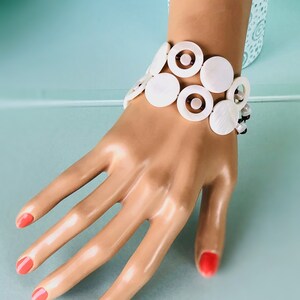 May include: White shell bracelet with brown accents. The bracelet is made of multiple layers of large, round shells with a smaller, inner circle of brown shell.