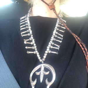 Puede incluir: Un collar de plata con un gran colgante adornado en forma de herradura. El collar está hecho de pequeñas cuentas de plata y tiene una franja de cuentas de plata que cuelgan de él.