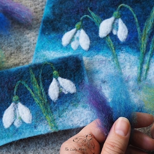 Könnte beinhalten: Zwei kleine Nadelfilzbilder von Schneeglöckchen auf blauem Hintergrund. Die Bilder befinden sich auf einer grauen Oberfläche mit Wollfasern im Vordergrund. Der Text "The Lady Moth" ist in der rechten unteren Ecke sichtbar.