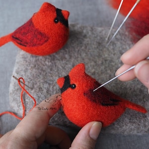 Op de afbeelding: Twee rode viltkardinaalvogels met zwarte ogen en zwarte markeringen op hun vleugels worden met een naald geviltd. De vogels worden in de handen van een persoon gehouden. De persoon gebruikt een naaldviltgereedschap om details aan de vogels toe te voegen.