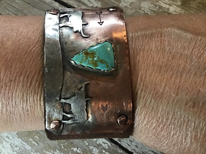 May include: A copper and silver cuff bracelet with a turquoise stone and arrow designs. The bracelet is worn on a person's wrist.