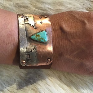 May include: A copper cuff bracelet with a turquoise stone and silver accents. The bracelet features a design of two stylized cattle and arrows.